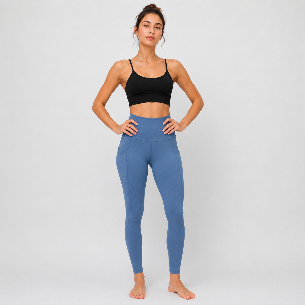 A woman stands barefoot against a light gray studio background wearing a black sports bra and high-waisted blue leggings.