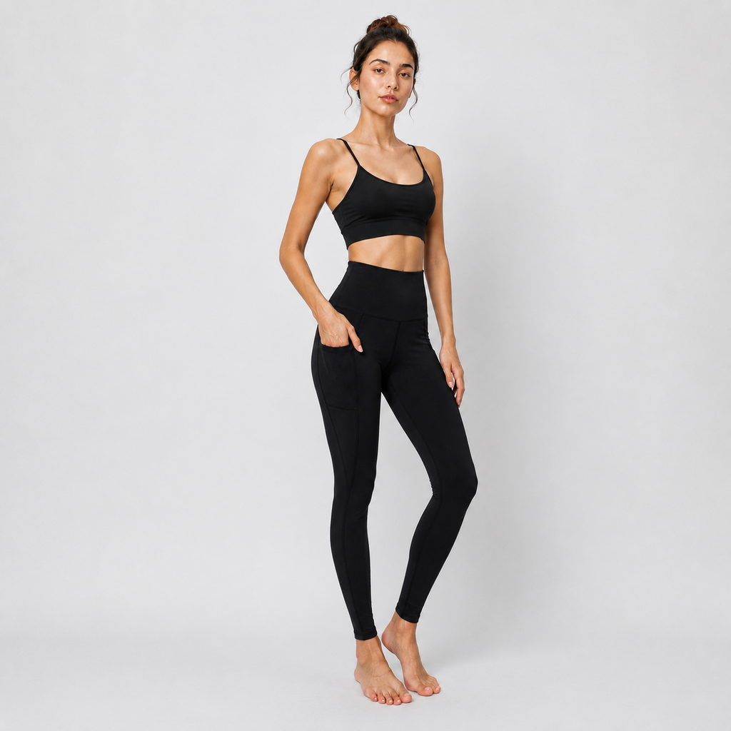 A woman modeling a black activewear set consisting of a sports bra and high-waisted leggings with side pockets, standing against a light gray studio background.