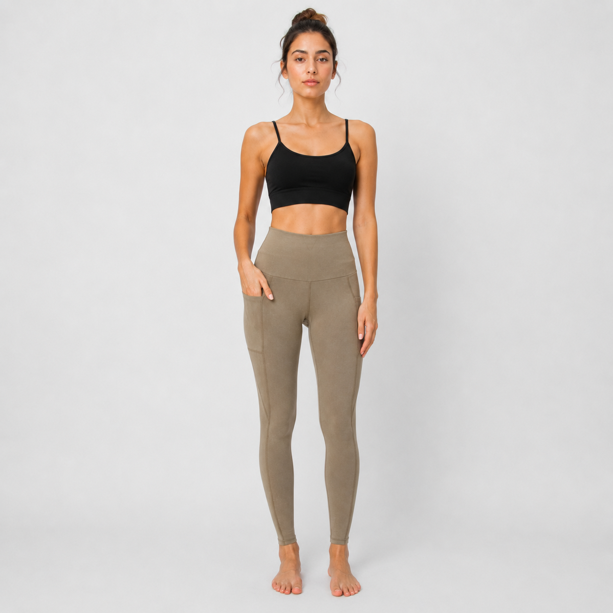 A woman stands barefoot against a light gray studio background wearing a black sports bra and high-waisted, side-pocket activewear leggings in a muted taupe color.