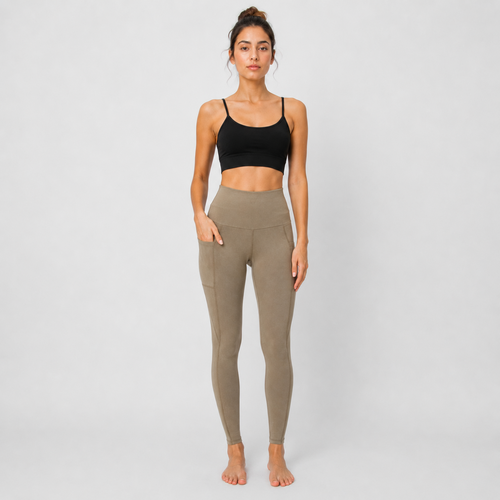 A woman stands barefoot against a light gray studio background wearing a black sports bra and high-waisted, side-pocket activewear leggings in a muted taupe color.