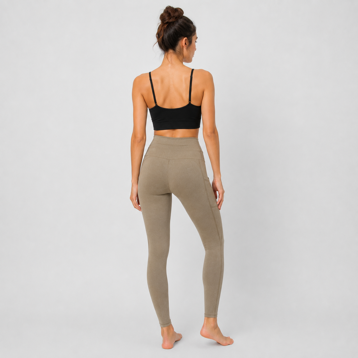 A woman standing with her back to the camera wearing a black sports bra and high-waisted taupe leggings against a light gray studio background.