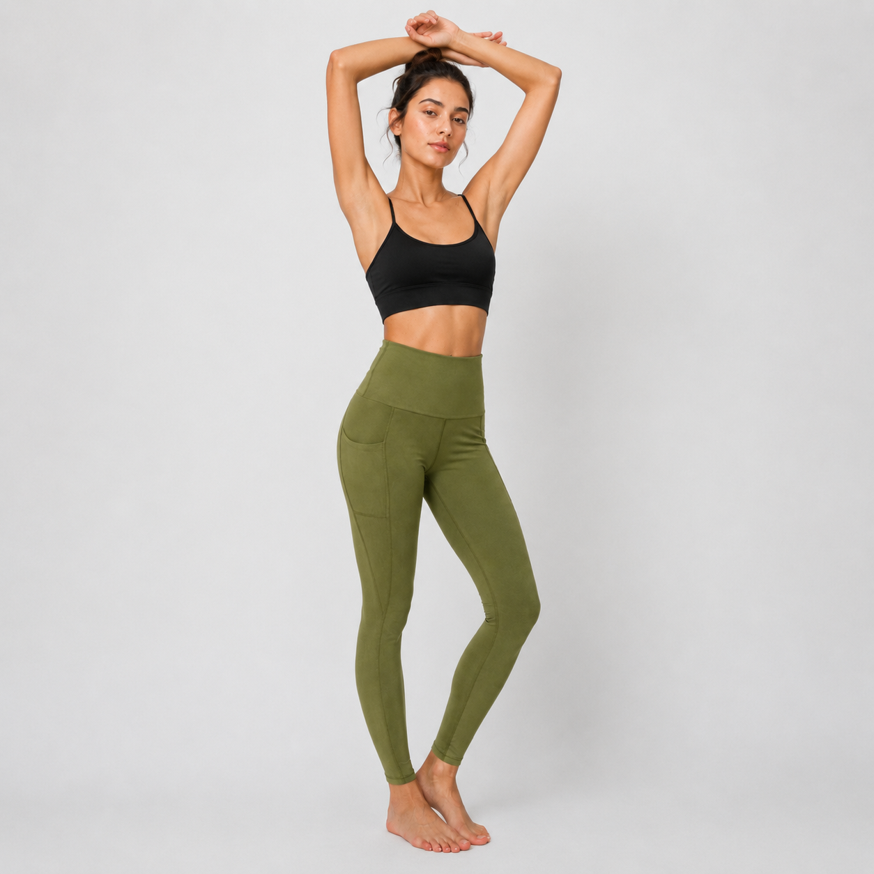 A woman modeling high-waisted olive green leggings and a black sports bra against a light gray studio background.